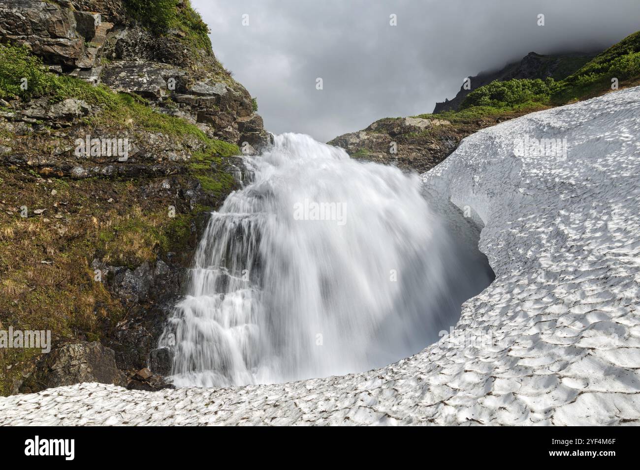 Incredibile paesaggio montano estivo della penisola di Kamchatka: Splendida vista della tranquilla cascata che cade nel campo innevato della catena montuosa Vachkazhet Foto Stock