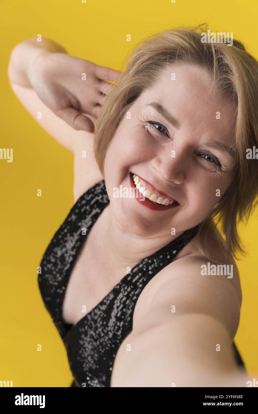 Ritratto di una donna bionda adulta sorridente e felice in un abito da festa nero con paillettes e scollatura che si allunga la mano in avanti per scattare un selfie e un aspetto giocoso Foto Stock