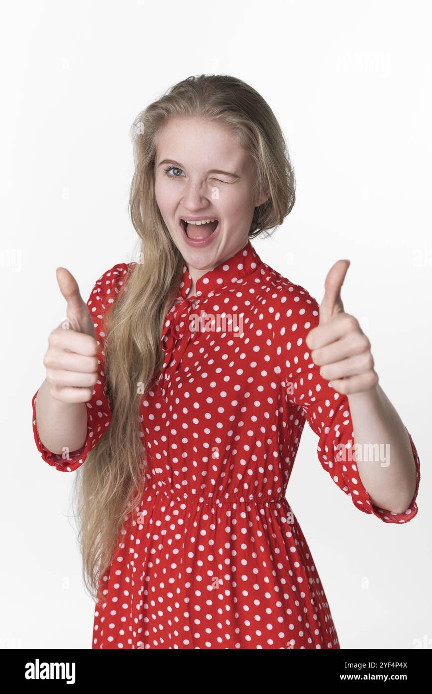 La felicità bionda ragazza ha chiuso giocosamente un occhio e mostra i pollici in alto con entrambe le mani. Ritratto di una giovane donna adulta sorridente vestita d'estate Foto Stock