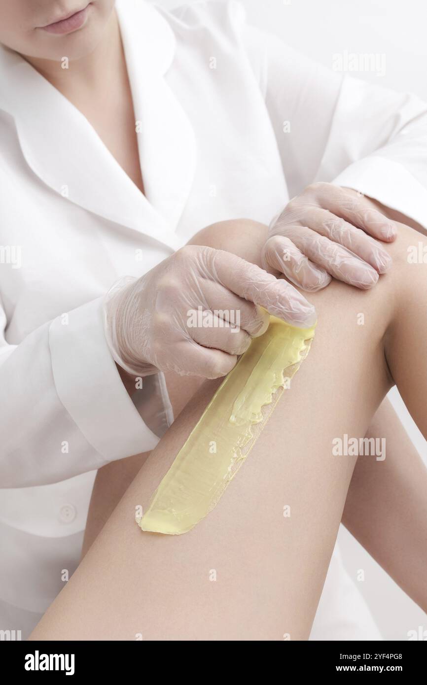 Vista di primo piano del cosmetologist che rimuove i capelli sulle gambe delle donne usando la cera calda. Rapido movimento improvviso di stoffa con cera calda verde in salone di bellezza professionale Foto Stock