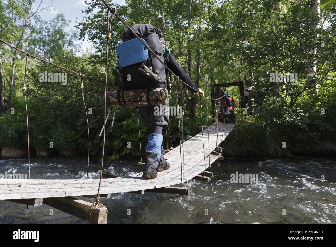 Escursioni estive sulla penisola di Kamchatka: Turista e viaggiatore con zaino dietro le spalle che attraversa il fiume di montagna nella foresta su suspensi pedonali Foto Stock