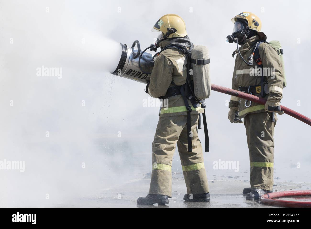 Due vigili del fuoco spengono il fuoco dal tubo antincendio, utilizzando un cilindro in schiuma d'acqua antincendio con schiuma meccanica ad aria. Giorno dei pompieri di festa professionale. Foto Stock