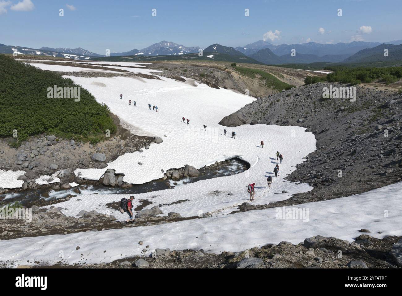 KAMCHATKA, RUSSIA, 10 LUGLIO 2014: I turisti attraversano il fiume di montagna camminando su campi innevati in estate giorno di sole. Russia, Estremo Oriente, penisola di Kamchatka Foto Stock