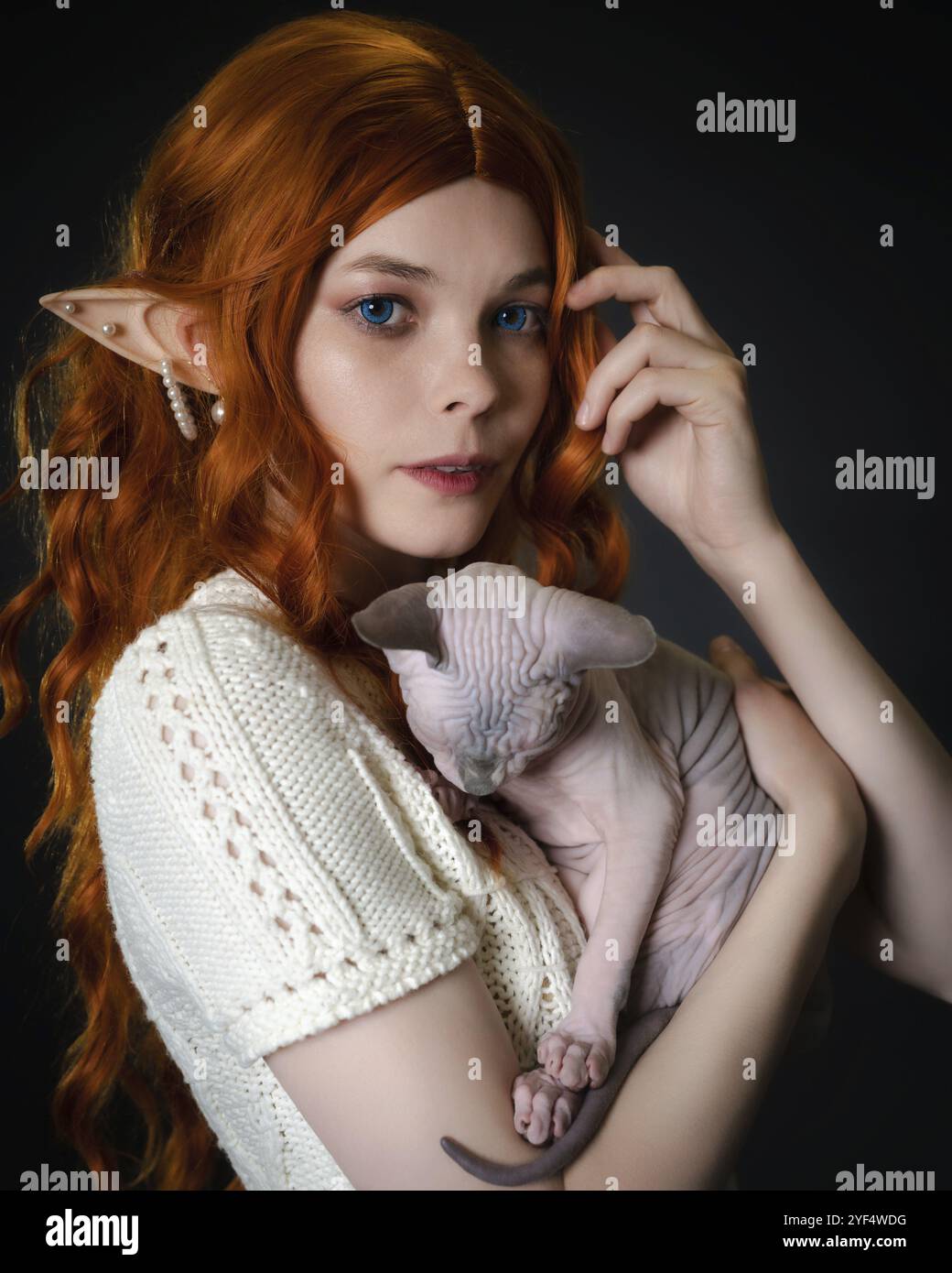 cosplay femminile giovane elfo con gattino Sfinge addormentato. Primo piano verticale su sfondo nero. Carino elfo in abito in maglia bianca, con capelli ricci rossi lunghi Foto Stock