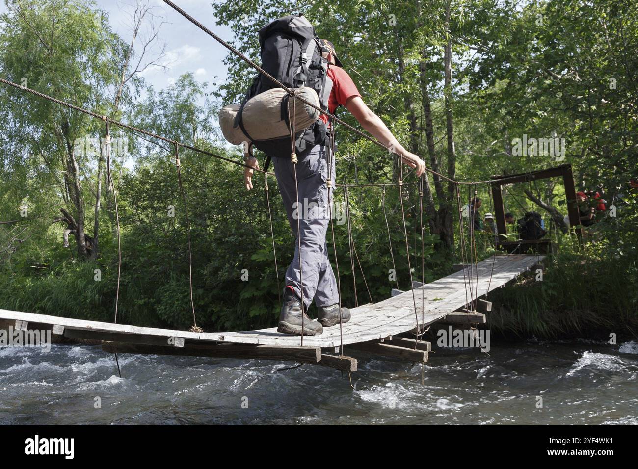 Tracciando sulla penisola di Kamchatka, escursionista con zaino dietro le spalle attraversando il fiume di montagna su un ponte sospeso pedonale nella foresta estiva. Come Foto Stock