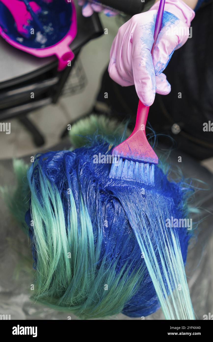 Ripresa ad alto angolo dei capelli di tintura di processo in salone di bellezza. Parrucchiere in guanto protettivo con spazzola magenta mentre si applica vernice blu alla donna con em Foto Stock