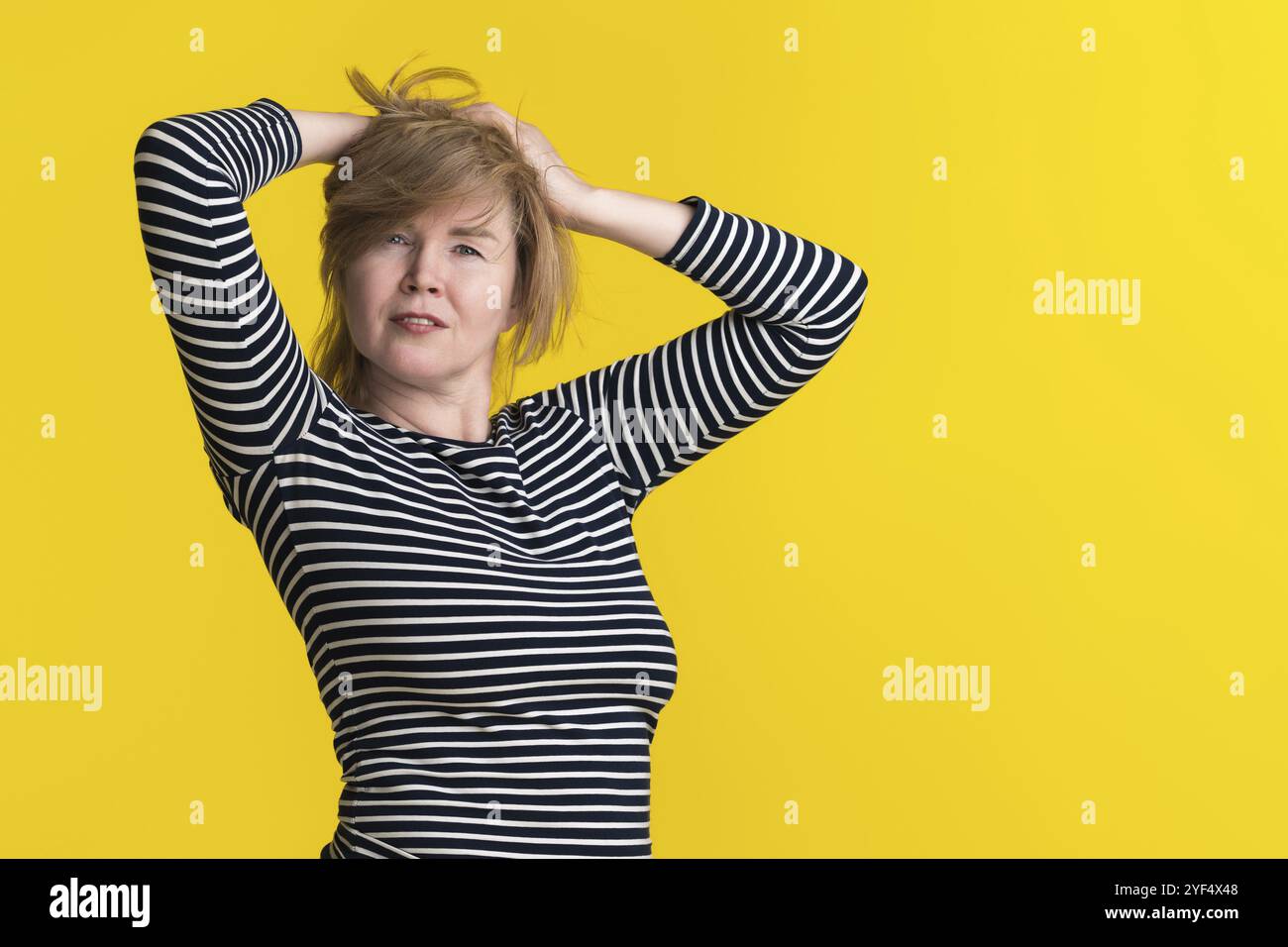 Una donna matura dai capelli biondi indossa una camicia a maniche lunghe a righe, solleva le mani dietro la testa e guarda la fotocamera. Lo sfondo è giallo solare Foto Stock