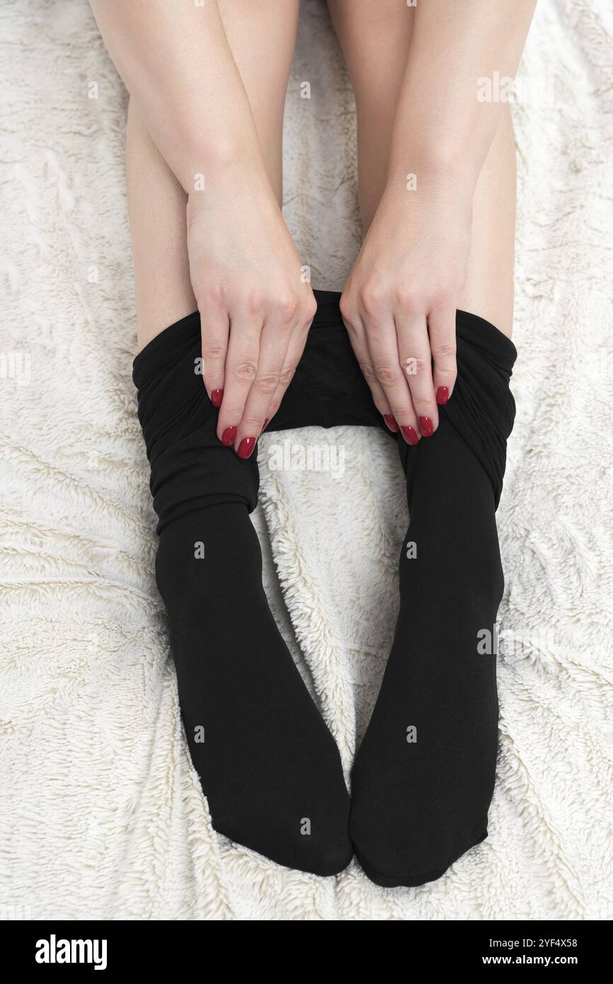 Vista ravvicinata delle mani femminili indossando con cura il collant nero. Vista rifilata delle timide gambe irriconoscibili della donna sul letto bianco. Vista dall'alto anteriore. Parte di S. Foto Stock