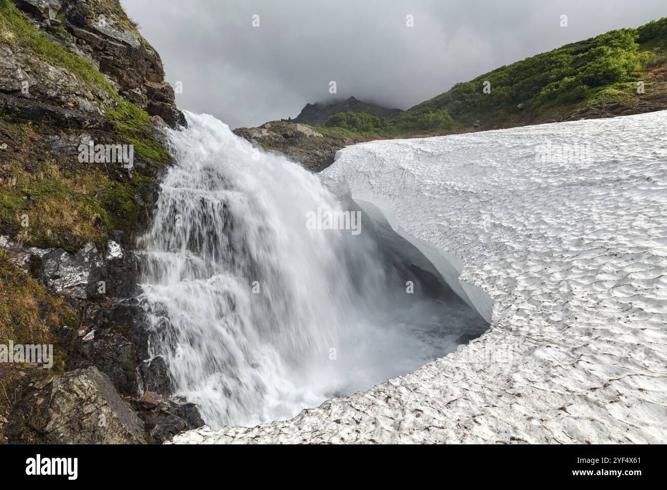 Incredibile paesaggio montano di Kamchatka: Vista estiva della splendida cascata che cade nel campo innevato della catena montuosa Vachkazhets. Eurasia, Russia Foto Stock