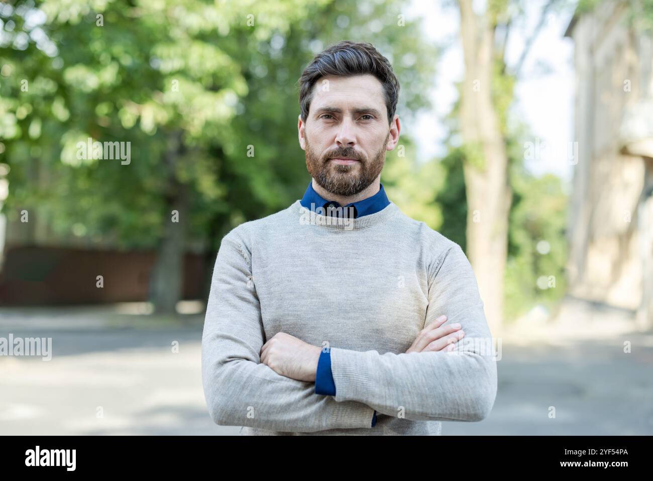 Ritratto di uomo maturo e sicuro di sé con le braccia incrociate in piedi all'aperto. Esprime autosicurezza e autorevolezza sullo sfondo sfocato di alberi ed edifici. Comportamento informale e aggressivo. Foto Stock