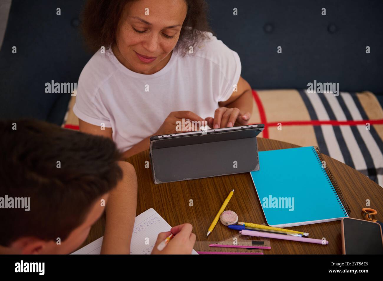 Una donna e un bambino impegnati nell'apprendimento con un tablet, blocchi appunti e cancelleria su un tavolo di legno, incoraggiando un'esperienza educativa condivisa in un ambiente accogliente Foto Stock