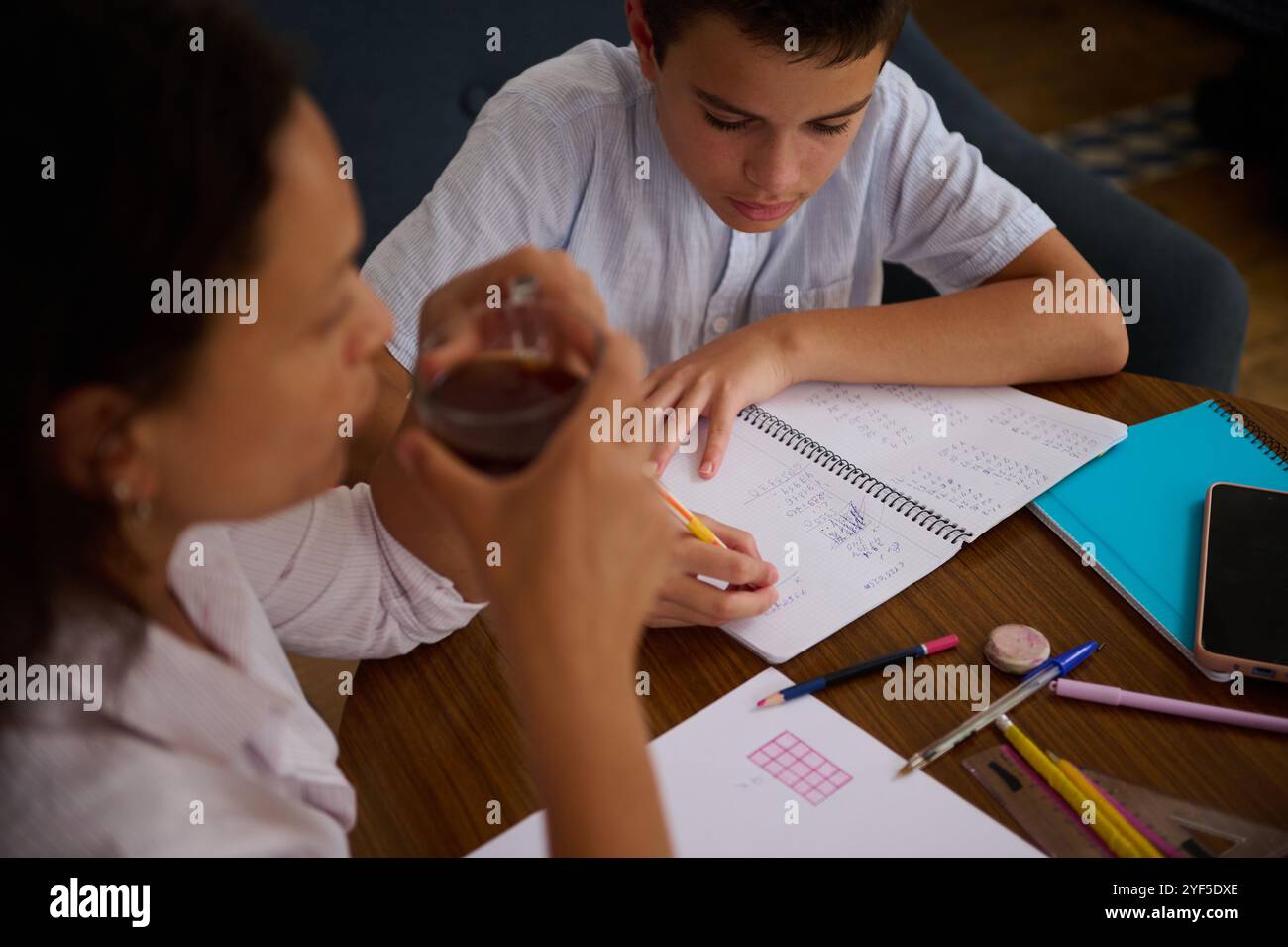 Un ragazzino si concentra sui compiti di matematica, tirando giù appunti in un quaderno a spirale. Un adulto, possibilmente un genitore o un tutor, si siede nelle vicinanze, fornendo la Foto Stock