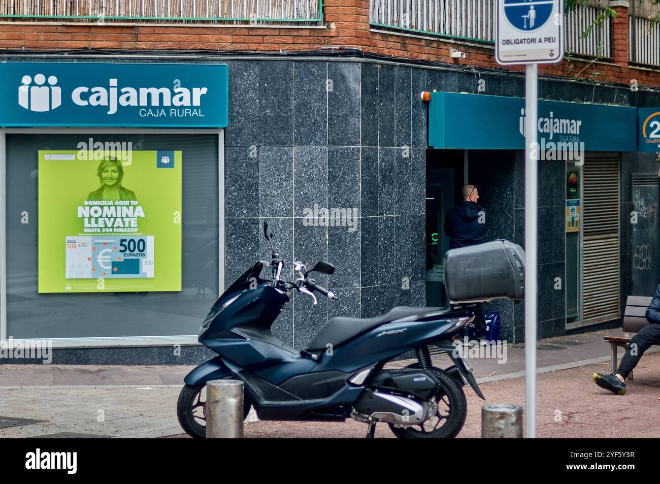 Viladecans. Spagna - 3 novembre 2024: Un moderno scooter nero è parcheggiato fuori da una banca rurale di nome Cajamar con un poster promozionale nella finestra e. Foto Stock