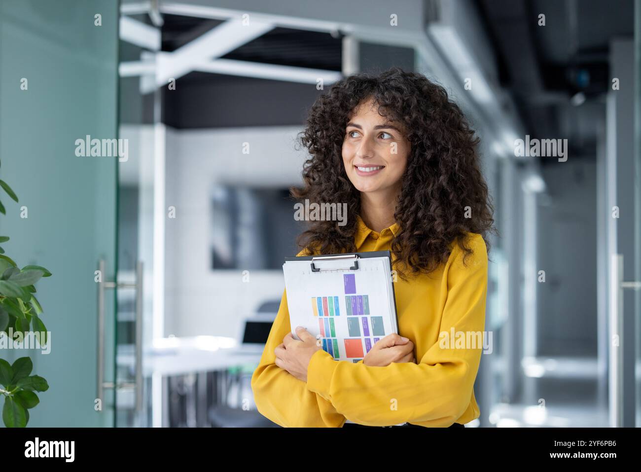 Donna d'affari sicura con capelli ricci in un ufficio moderno contiene appunti con grafici colorati. Irradia ispirazione e attenzione in un ambiente professionale. Foto Stock