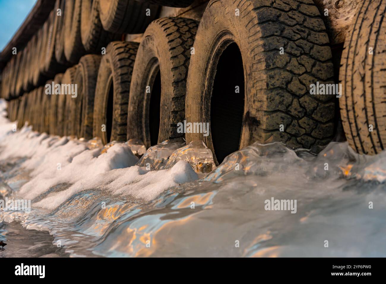 Grandi blocchi di ghiaccio ghiacciati su un porticciolo con pneumatici per auto in inverno. Il tramonto dorato si illumina sul lato di un lago ghiacciato in inverno. Foto Stock