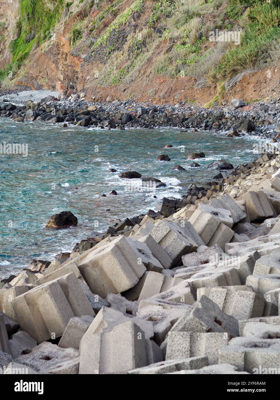 L'aspra linea costiera è caratterizzata da blocchi esagonali di cemento sparsi lungo la costa, adagiati su un mare blu abbagliante con dolci onde che toccano la roc Foto Stock