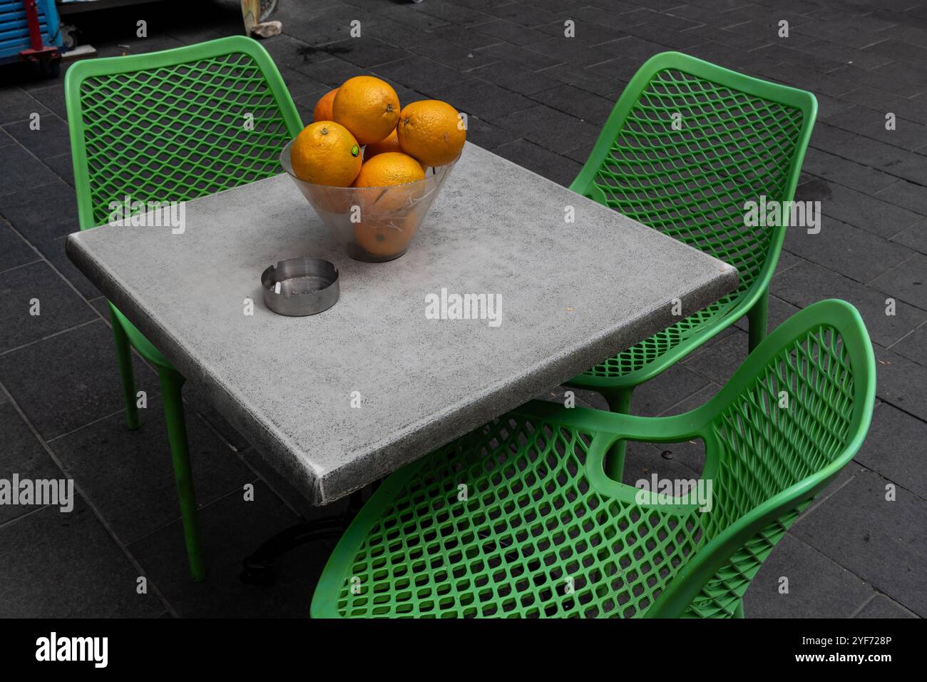 Immagine minimalista di una ciotola di arance su un tavolo quadrato circondato da tre sedie verdi di plastica su un marciapiede di Gerusalemme. Foto Stock
