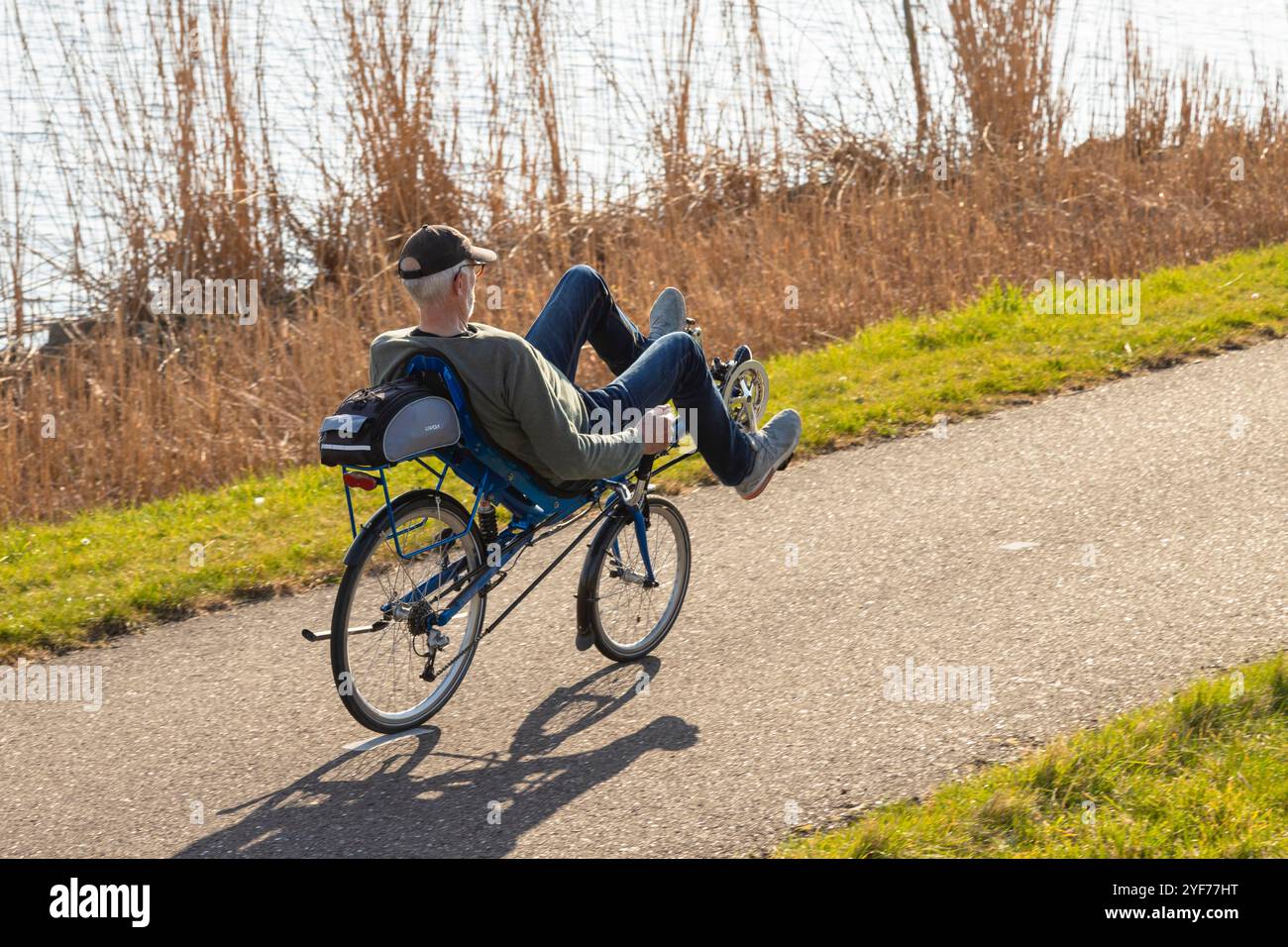 Un uomo anziano guida una bicicletta reclinata lungo l'acqua con le canne. Foto Stock