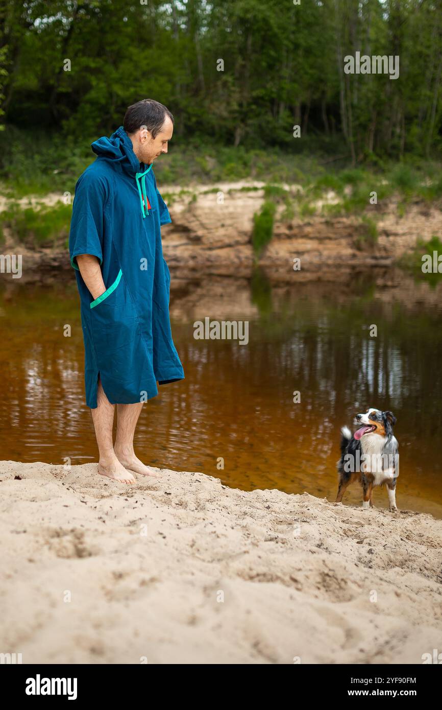 Man in Teal Towel Poncho posa vicino al fiume, Watchful Dog si trova nelle vicinanze Foto Stock