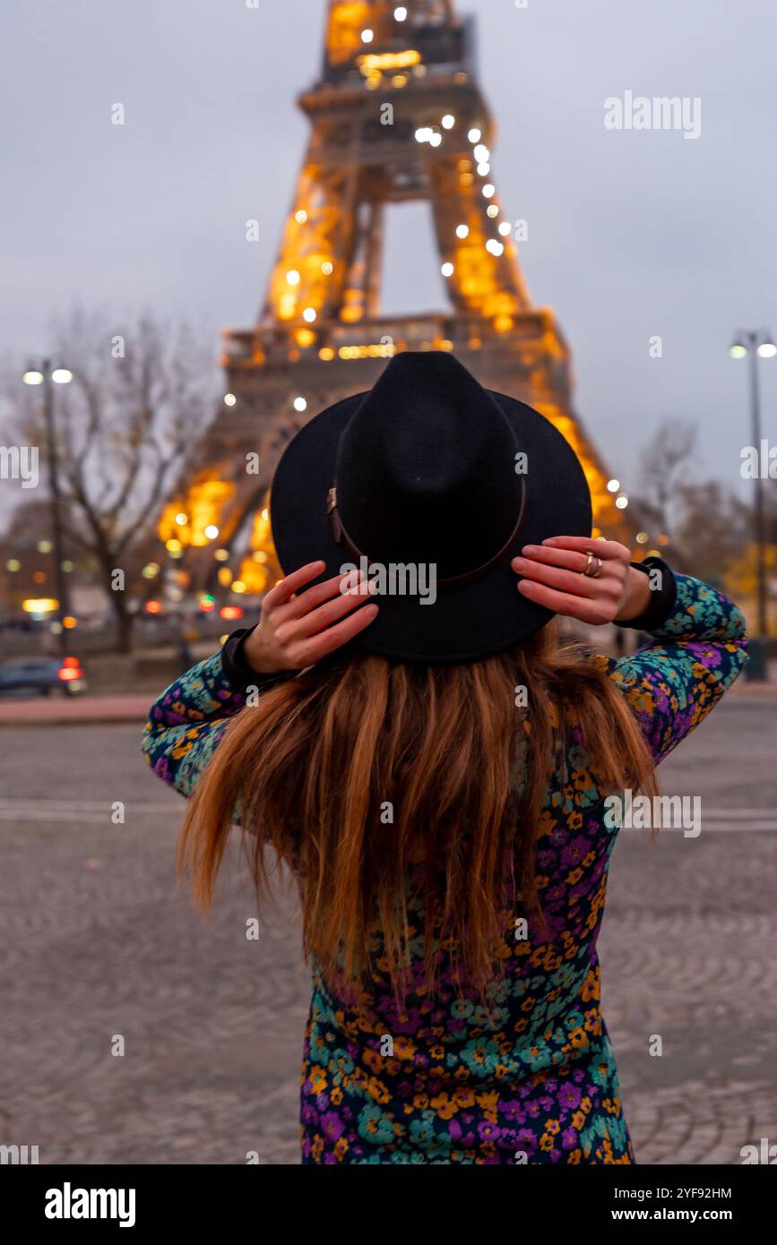 Donna alla moda con abito floreale e cappello in posa con la Torre Eiffel illuminata al tramonto, l'eleganza parigina incontra l'iconico simbolo francese Foto Stock