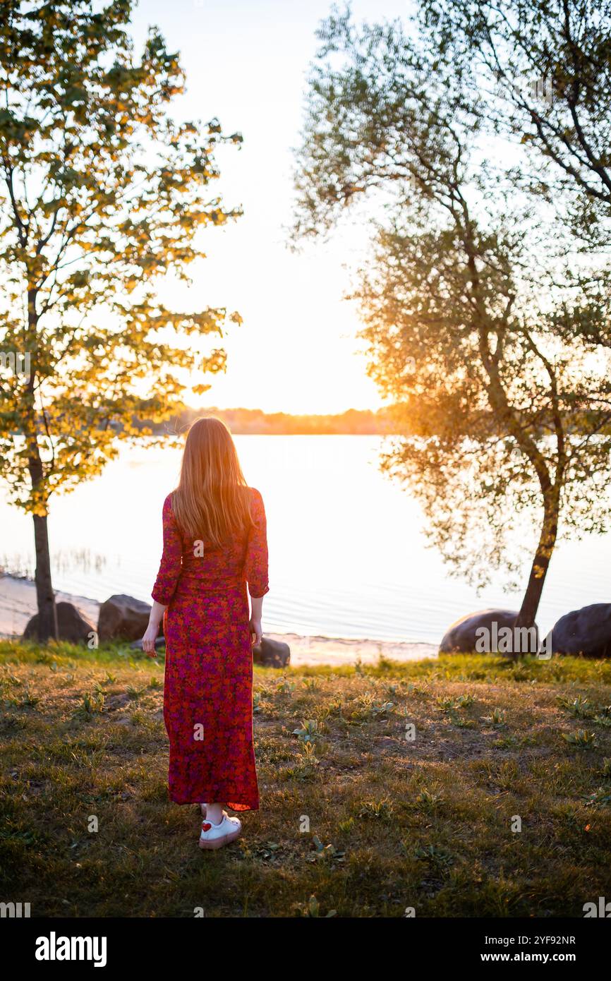 donna in un abito che scorre ammirando il tramonto in riva al lago, un momento di solitudine avvolto dal calore della luce dorata Foto Stock
