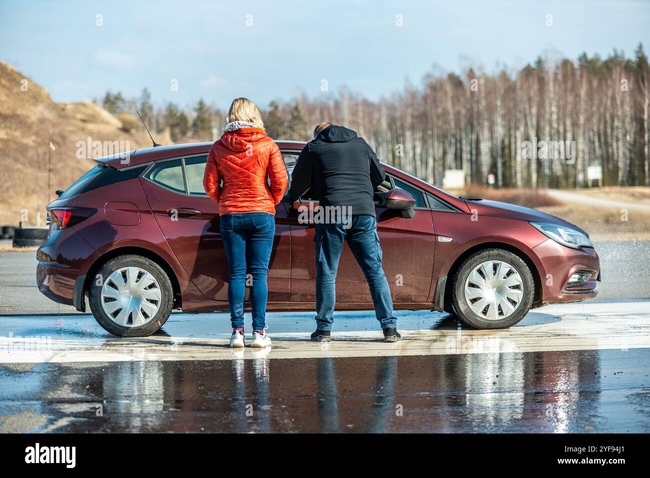 uomo che osserva una prova su strada bagnata con un'auto color marrone che slitta su una pista coperta d'acqua, prova la sicurezza del veicolo e le prestazioni degli pneumatici sotto controllo Foto Stock