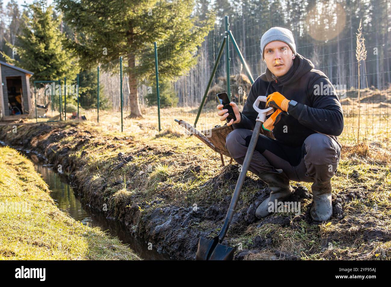 Uomo che fa una pausa dal lavoro all'aperto per controllare il telefono mentre si inginocchia davanti a un fosso di drenaggio scavato di recente in un giardino lussureggiante. Foto Stock