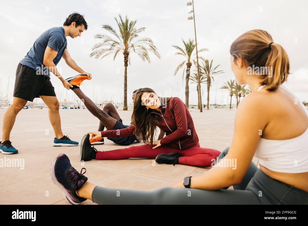 Un gruppo di amici partecipa a una sessione di fitness stretching all'aperto, divertendosi insieme in riva al mare con le palme sullo sfondo. Foto Stock