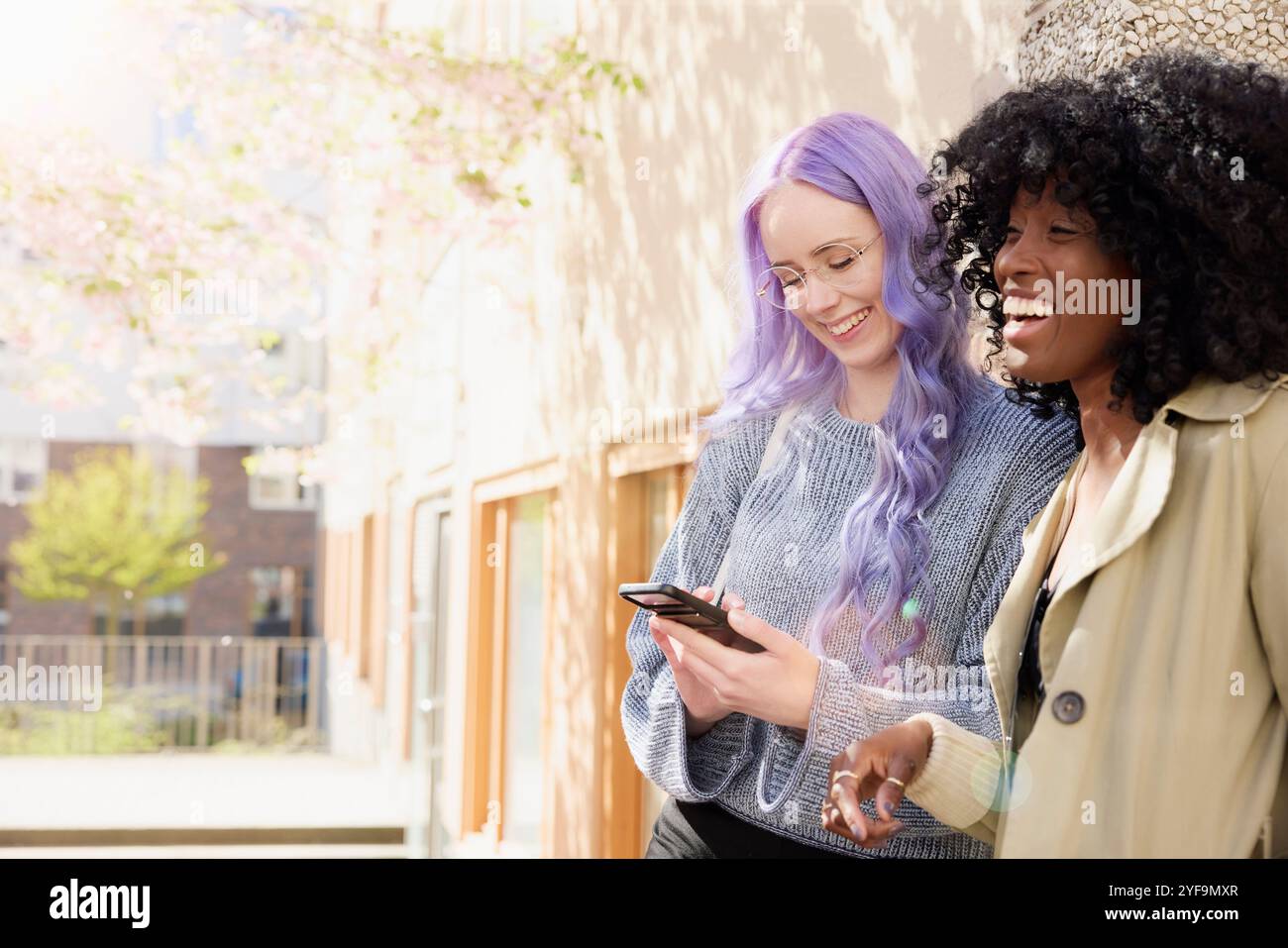 Donna felice con i capelli viola che utilizza lo smartphone e che si diverte a divertirsi con un'amica in strada Foto Stock