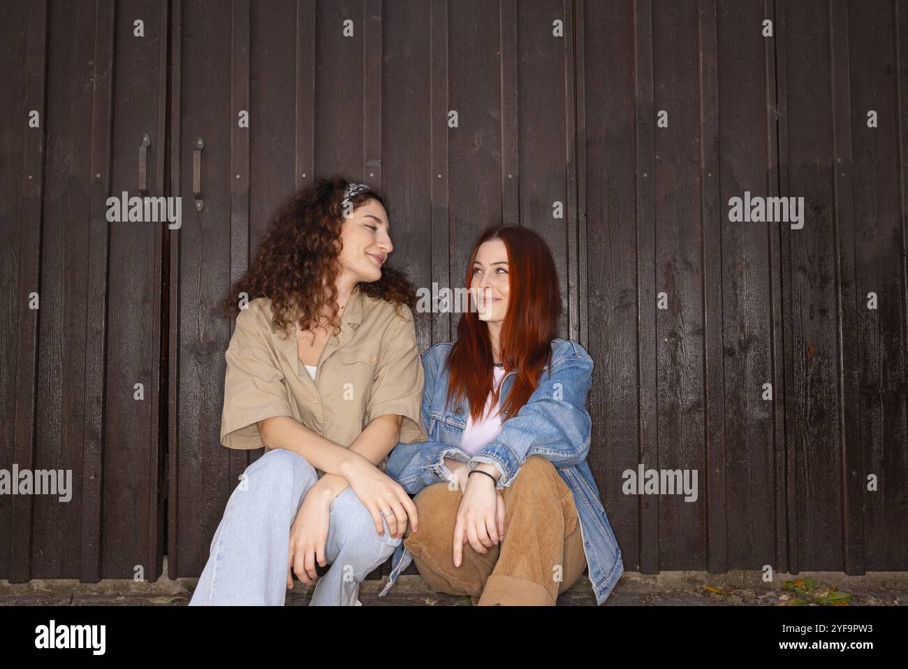 Amiche donne che si guardano mentre si siedono contro il cancello marrone Foto Stock