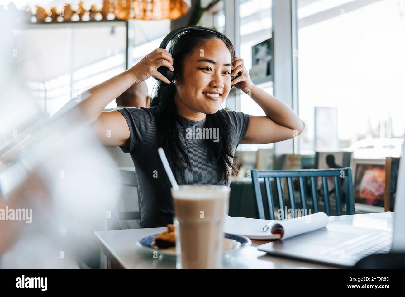 Giovane donna sorridente che indossa le cuffie mentre è seduta in un bar Foto Stock