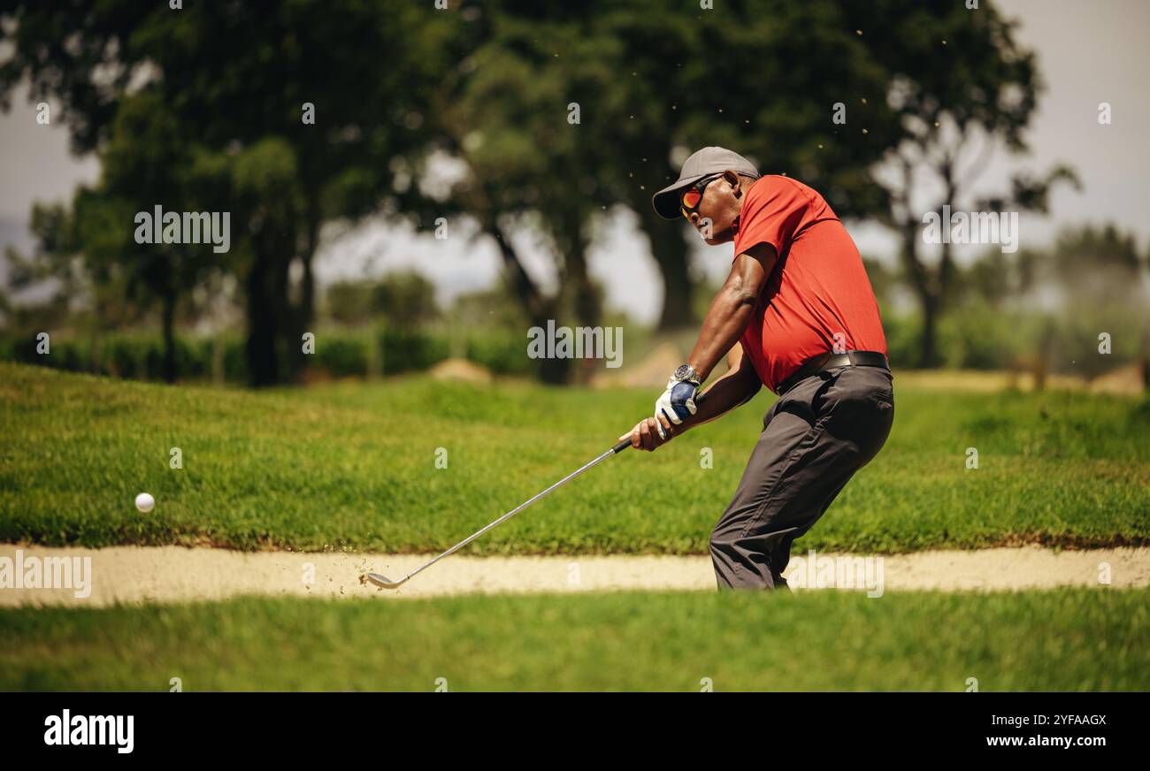 Uomo concentrato che gioca a golf in un bunker di sabbia, colpendo la palla con precisione. Cattura l'intensità e la concentrazione del gioco. Foto Stock