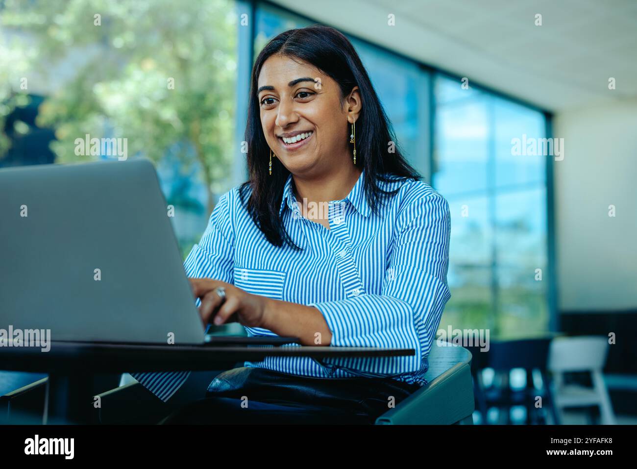 Una donna d'affari indiana è impegnata in una riunione virtuale, sorridendo mentre lavora sul suo portatile. L'ambiente luminoso e il suo comportamento positivo suggeriscono Foto Stock