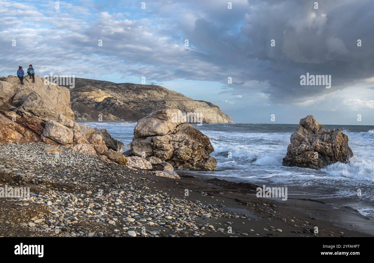 Due donne turistiche vestite con abiti invernali sedute su una roccia e godendosi lo splendido paesaggio marino tempestoso della spiaggia Rock of Aphrodite a Paphos, California Foto Stock