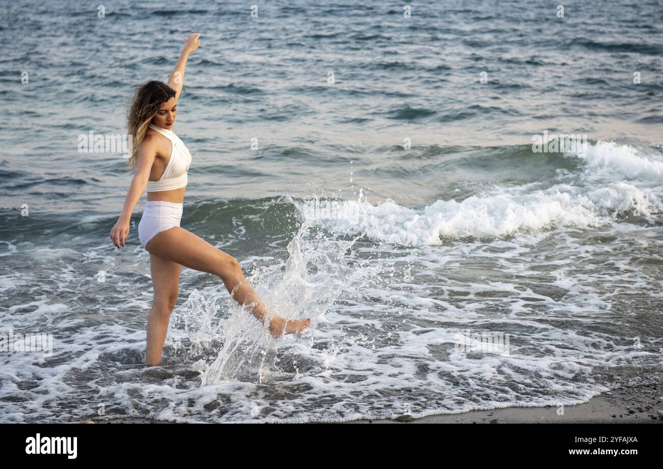 Giovane donna con costume da bagno bianco che gioca in acqua alla spiaggia al tramonto. Vacanze estive Foto Stock