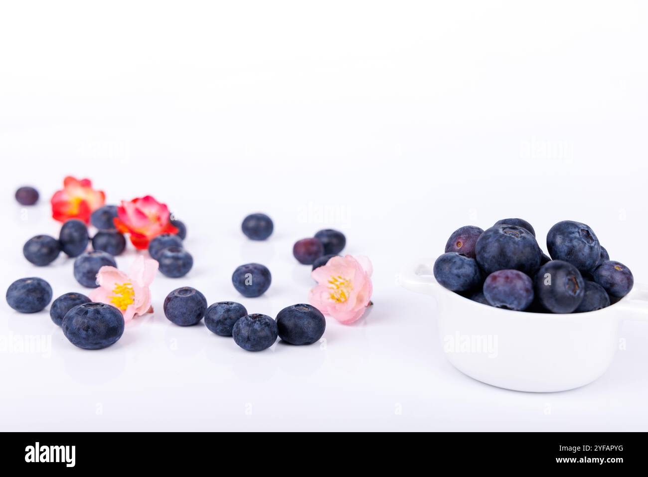 Una ciotola di mirtilli con fiori rossi e rosa sparsi su sfondo bianco, che mette in risalto freschezza, colore e semplicità in natura Foto Stock