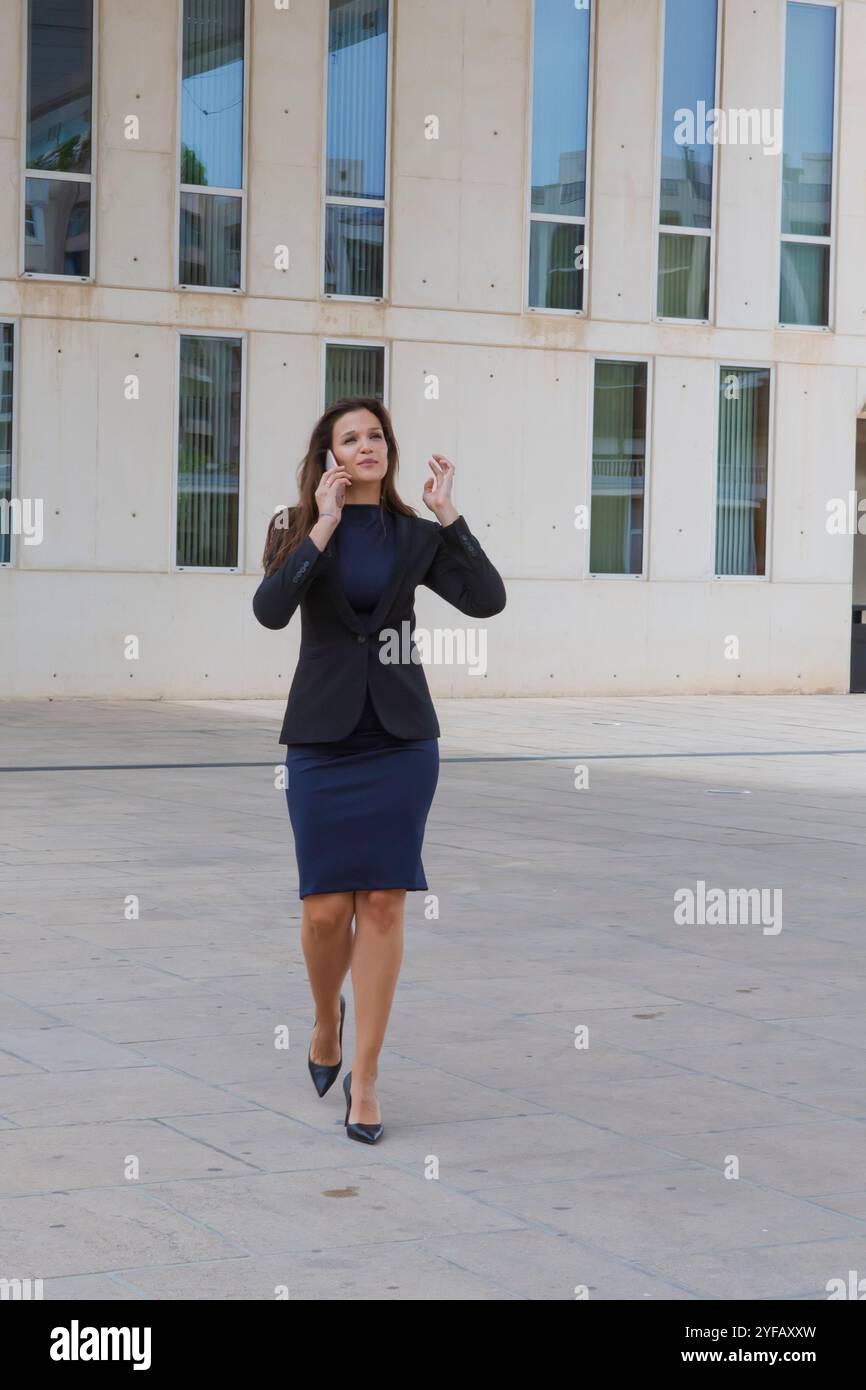 Donna professionista che cammina con sicurezza mentre parla al telefono cellulare in un ambiente urbano con architettura moderna sullo sfondo. Foto Stock