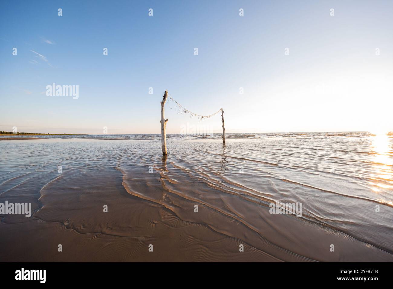 Primo piano di una spiaggia tranquilla con due pali di legno in piedi nell'acqua poco profonda, adornati con decorazioni a corda sotto la calda luce dell'alba. Foto Stock