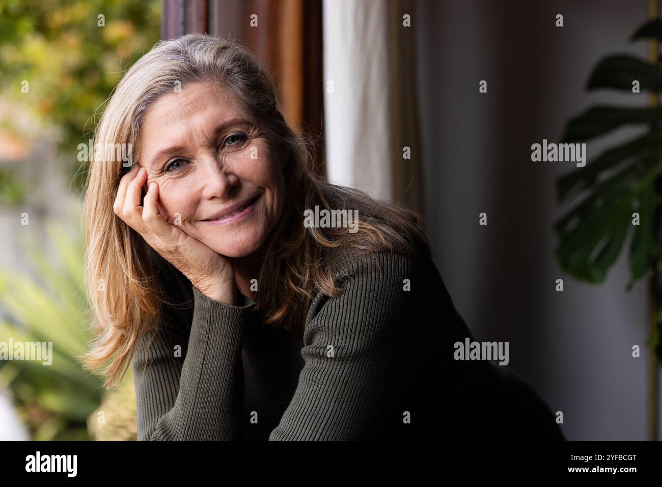 Donna anziana sorridente che gode di un momento tranquillo dalla finestra a casa Foto Stock