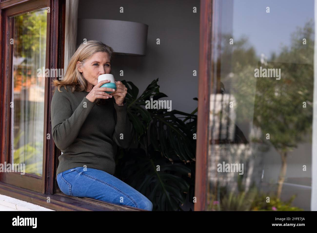 donna anziana che si rilassa a casa, beve un caffè vicino alla finestra, atmosfera tranquilla Foto Stock