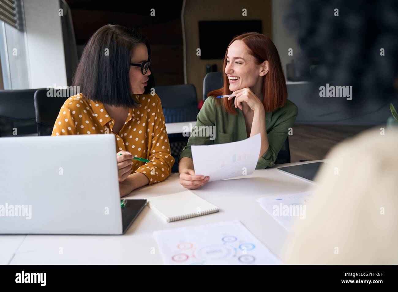 Due donne d'affari condividono una risata mentre analizzano grafici e dati durante una riunione produttiva in ufficio Foto Stock