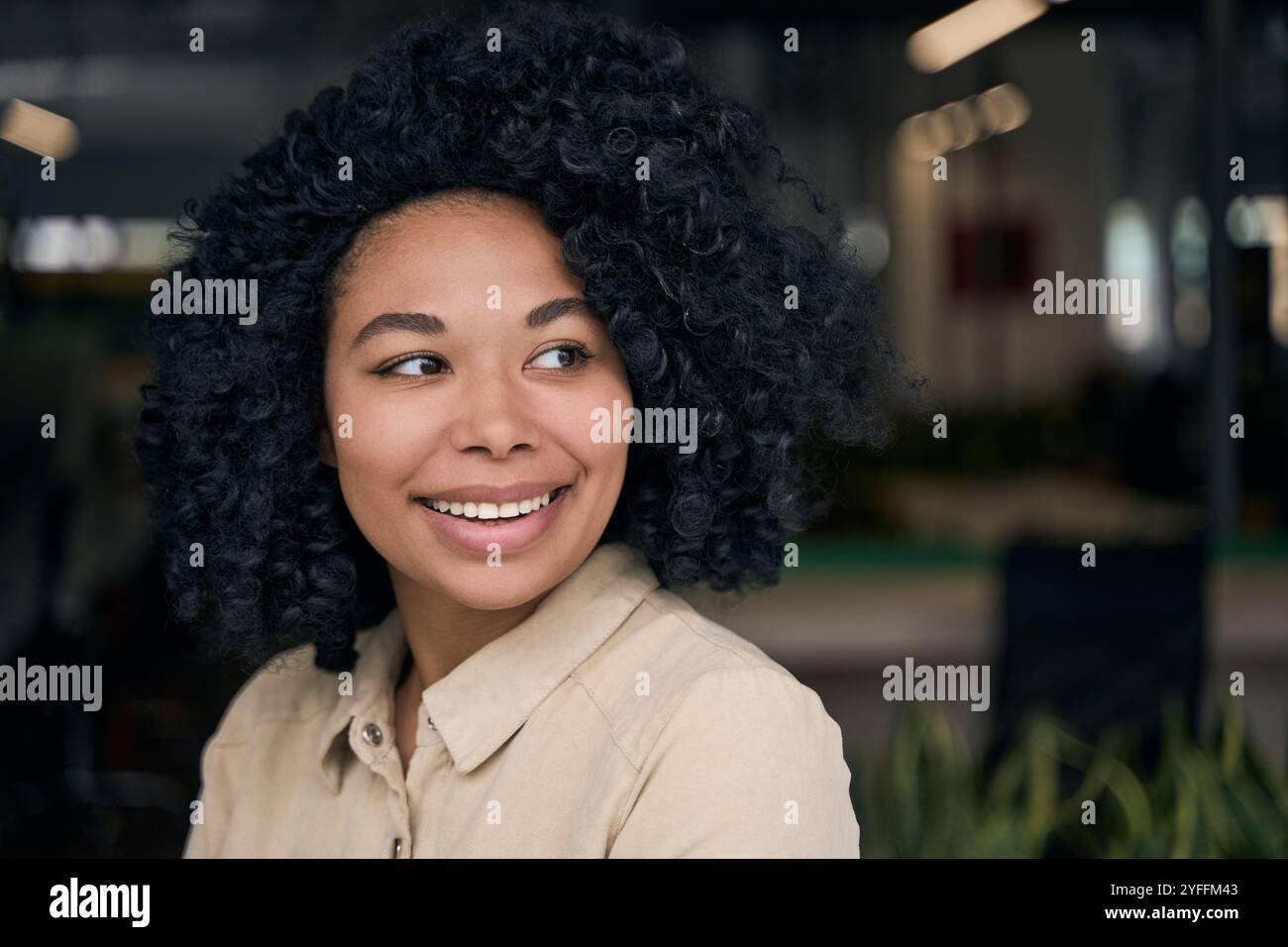 La giovane donna nera con i capelli ricci sorride mentre si guarda alle spalle in un ambiente d'ufficio Foto Stock