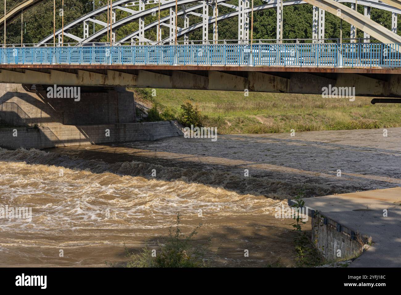 L'innalzamento delle acque fluviali fluiscono turbolentemente sotto un ponte d'acciaio arcuato durante la stagione delle inondazioni, evidenziando la potenza e l'imprevedibilità della natura Foto Stock
