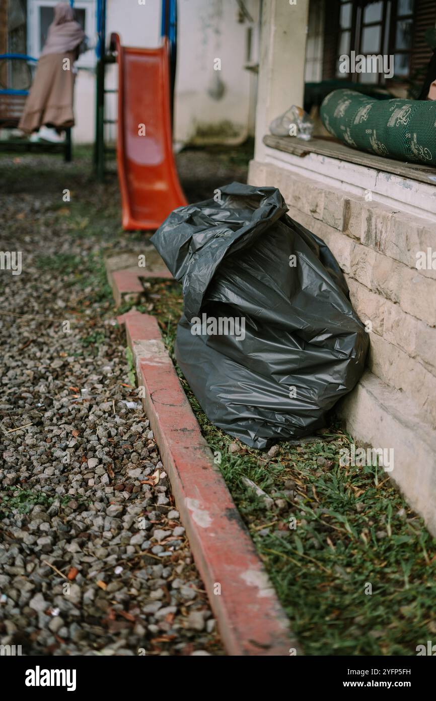 Un sacchetto nero per la spazzatura posto contro un muro di pietra, su un terreno coperto da piccoli ciottoli e macchie d'erba. Un cursore arancione è visibile sul retro Foto Stock