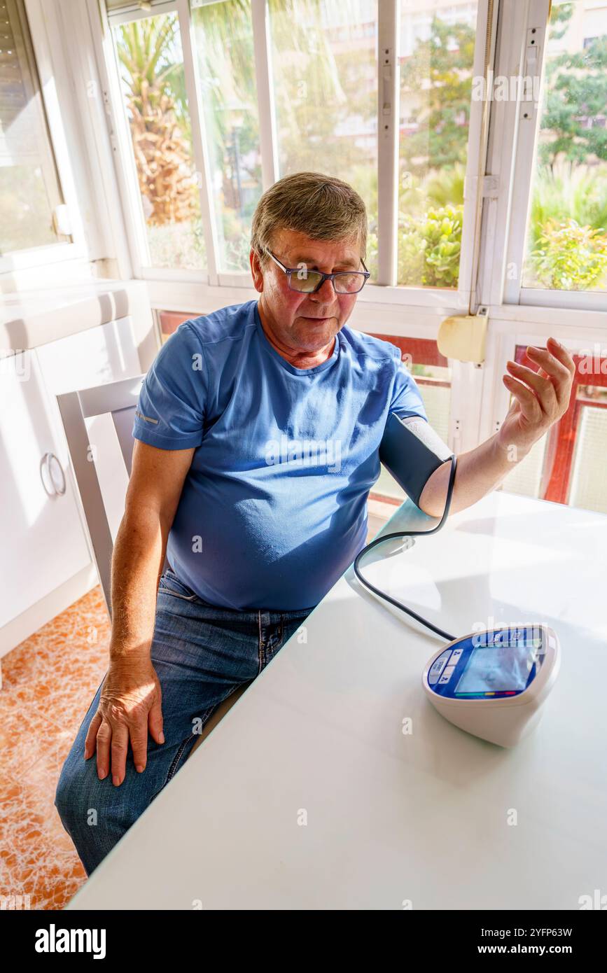 Uomo anziano in sovrappeso che misura la pressione sanguigna con il monitor a casa in soggiorno Foto Stock