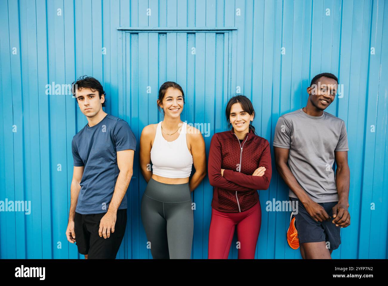 Ritratto di un gruppo di quattro amici di diverse origini che si appoggiano su una grande porta di metallo blu dopo una sessione di allenamento. Foto Stock