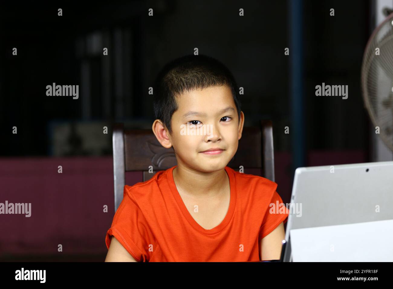 Un ragazzo allegro sorride mentre è seduto a una scrivania con un tablet, mostrando un'atmosfera gioiosa e giocosa in un ambiente interno, perfetto per l'istruzione e l'istruzione Foto Stock