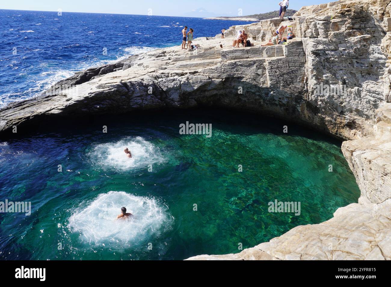 Questa foto mostra una splendida piscina naturale sul mare con acque turchesi cristalline circondata da rocce aspre e soleggiate. Due persone spruzzano in acqua. Foto Stock