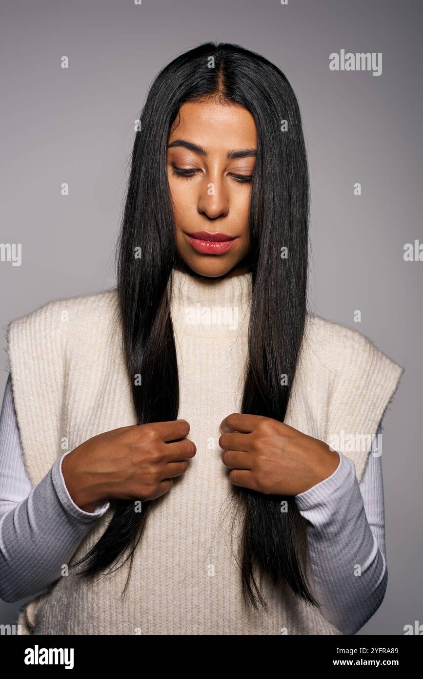 Una giovane donna con un maglione elegante tocca delicatamente i suoi lunghi capelli lisci chiudendo gli occhi. Foto Stock