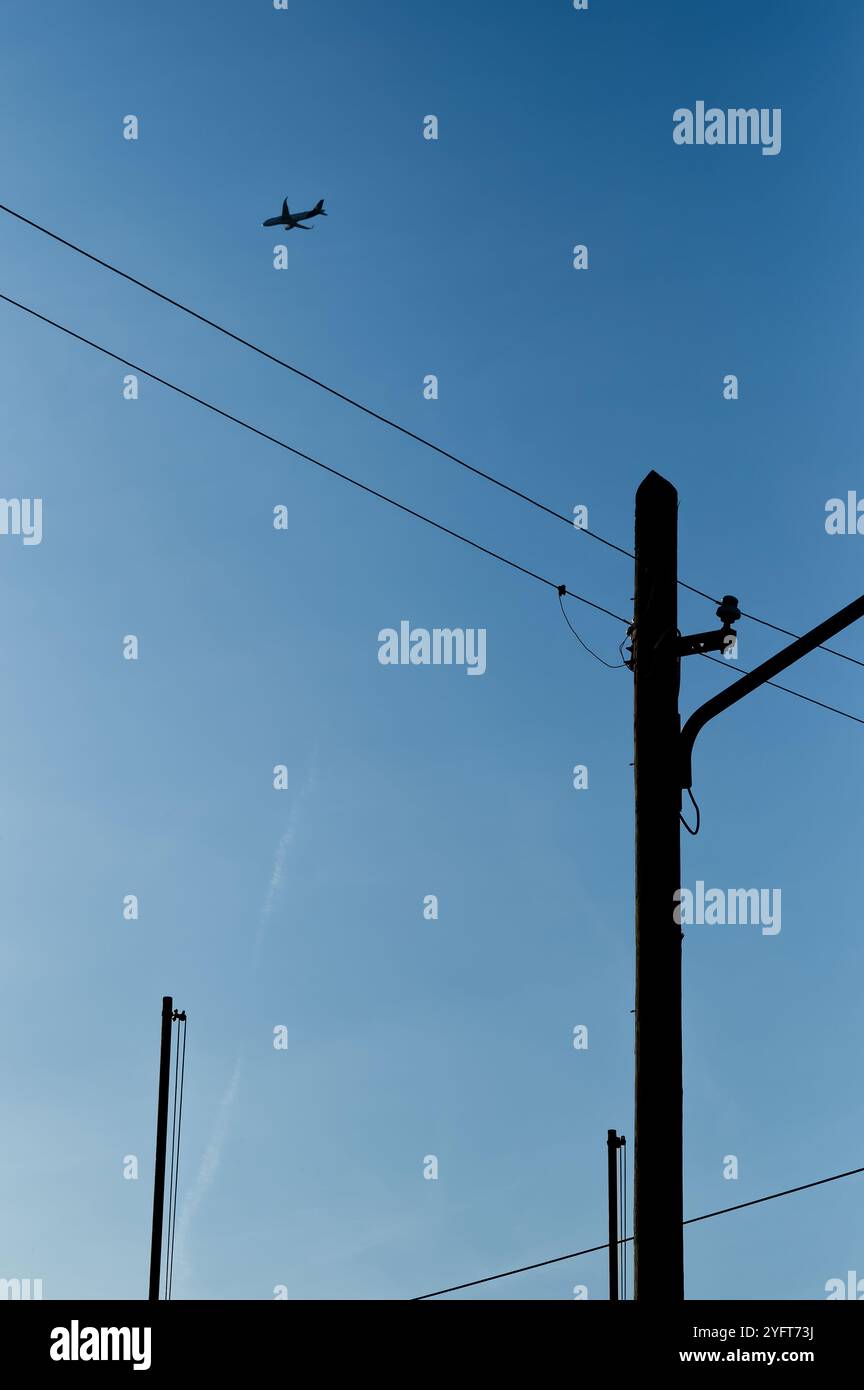 diversi pali di potenza e una doppia linea di alimentazione contro un cielo azzurro e un singolo aereo di linea nella parte superiore Foto Stock